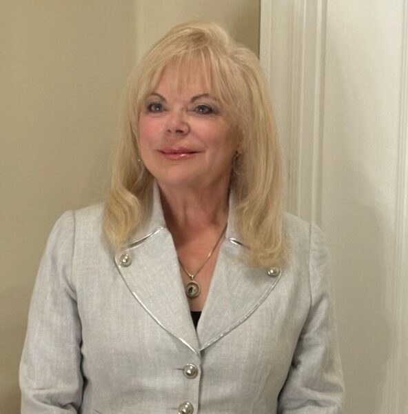 Teresa is a female with long blond hair and wearing a beige suit.